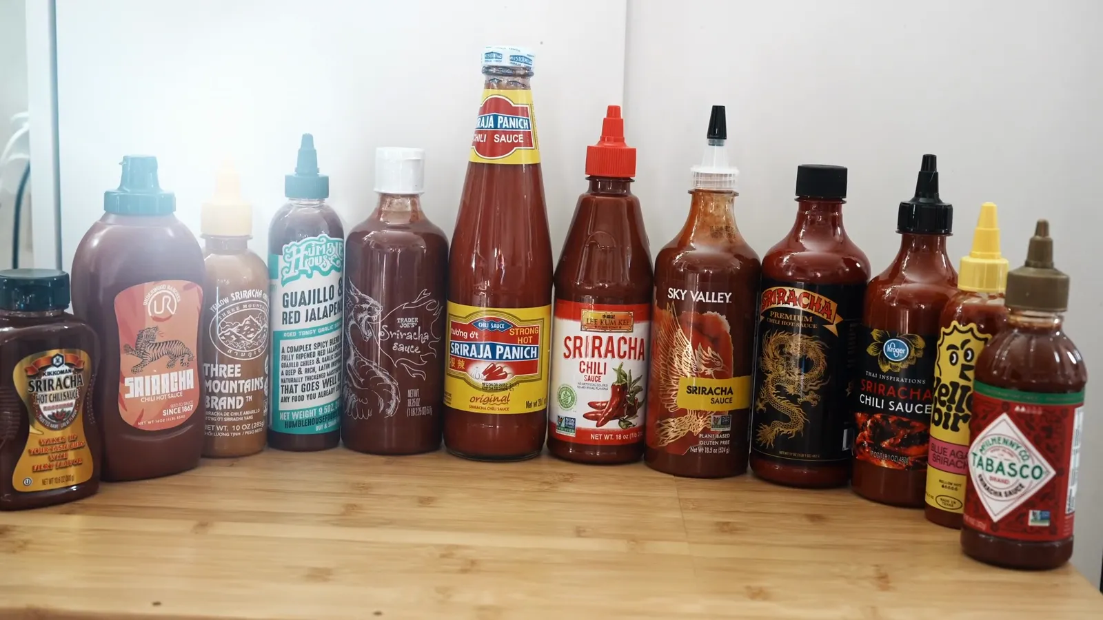 All 12 sriracha bottles we tested lined up on a counter: Kikkoman, Underwood Ranches, Three Mountains, Humble House, Trader Joe's, Sriraja Panich, Lee Kum Kee, Sky Valley, Underwood premium, Kroger, Yellowbird and Tabasco
